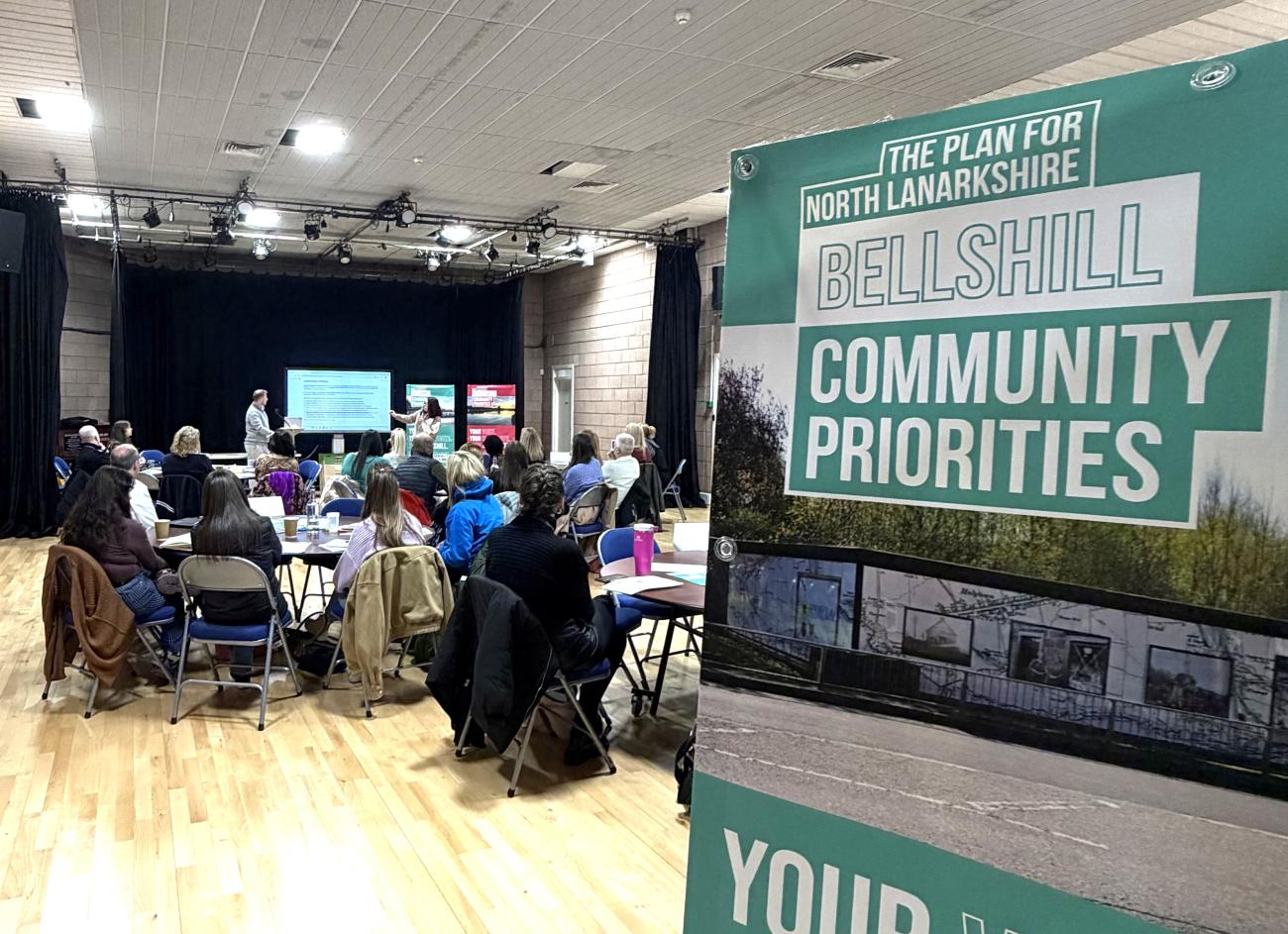 Bellshill community priority banner in focus with people seated in the background
