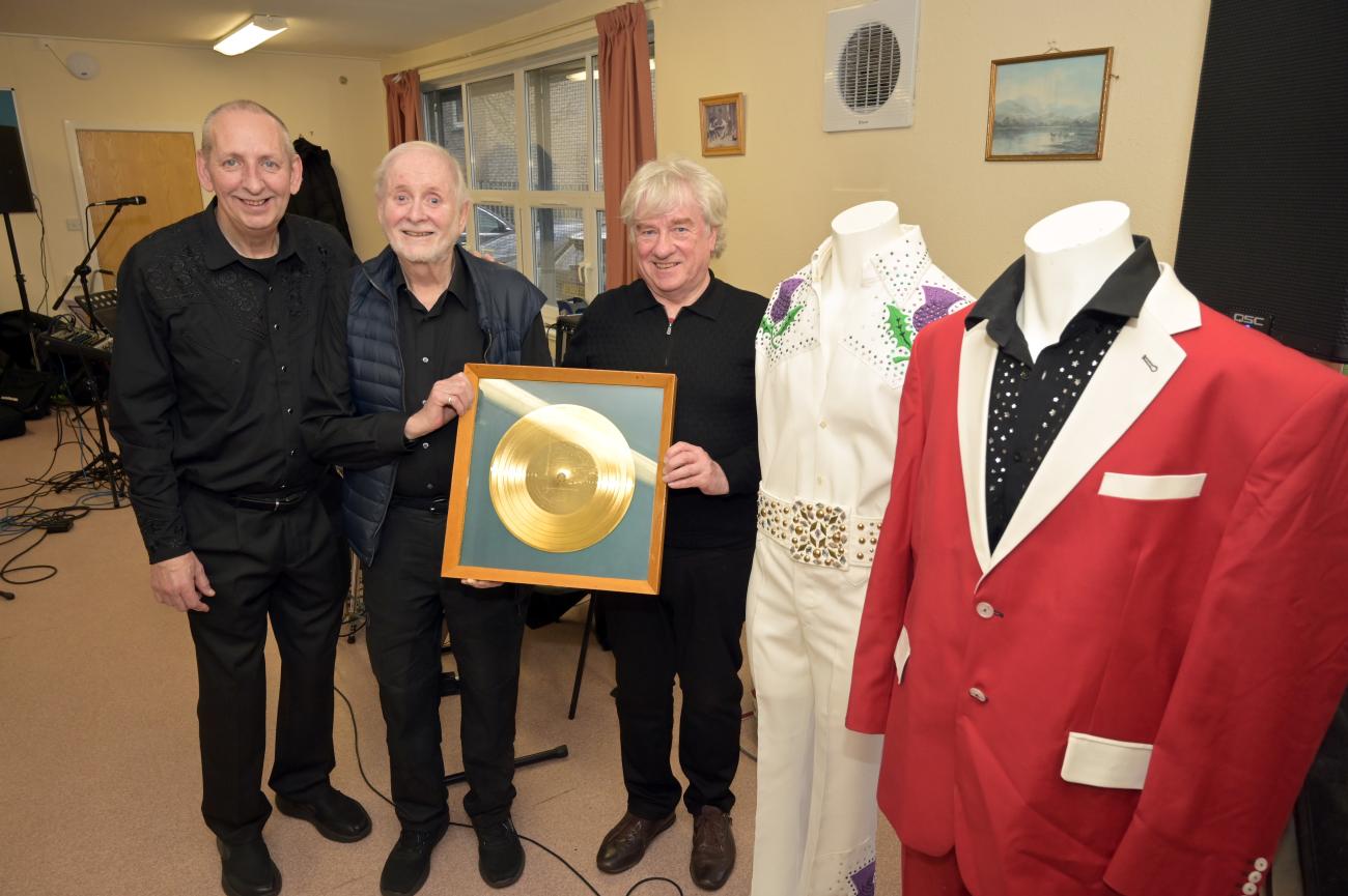 Sydney Devine's band members standing with his golden plaque and beside two of his famous outfits