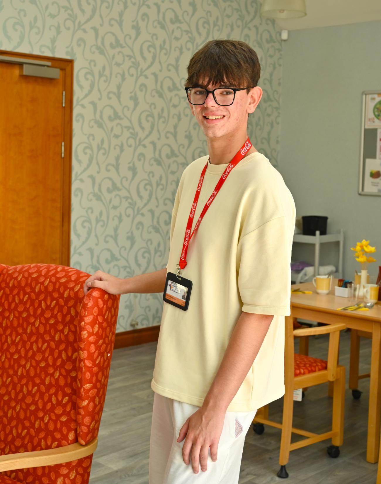 A Summer Placement wearing an ID badge stands in a care facility lounge beside several chairs, with dining tables and chairs visible in the background.