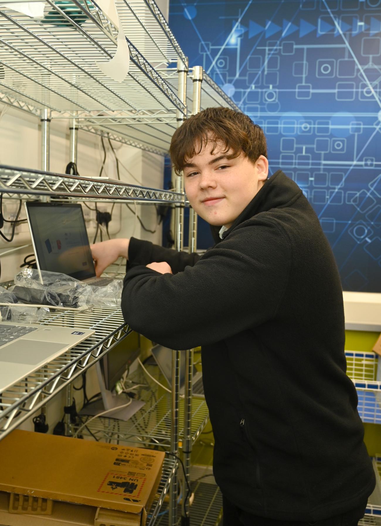 A Summer Placement works on a laptop at a workstation surrounded by computer equipment and shelving in a digital technology setting.