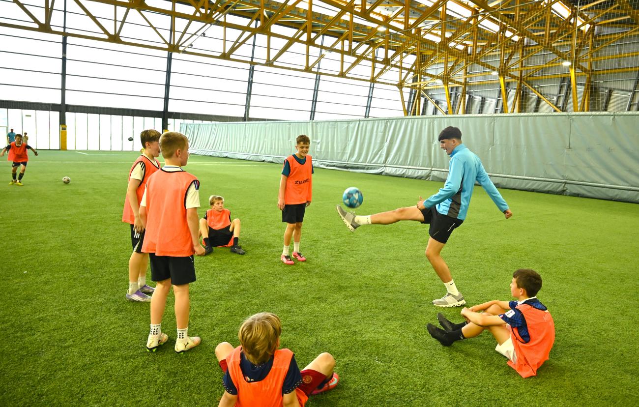 A Summer Placement participant leads a football activity for a group of young people on an indoor sports pitch.